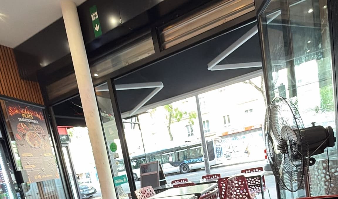 Salle du restaurant : tables noires, chaises rouges ajourées et grandes baies vitrées sur l’avenue